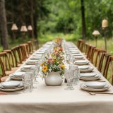 A long table with a white tablecloth and a vase of flowers in the center. The table is set with many plates, glasses, and utensils. Scene is elegant and inviting